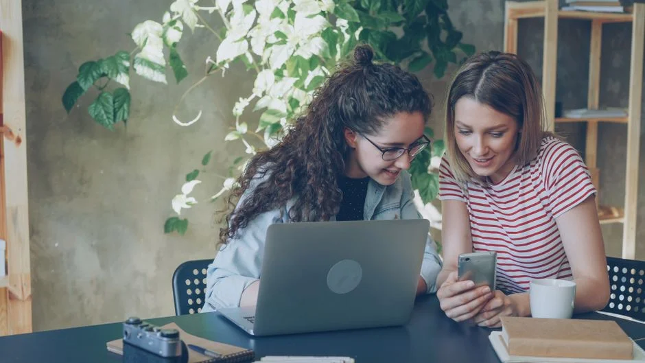 Zwei Kolleginnen schauen gemeinsam lächelnd auf ein Smartphone, im Hintergrund ein aufgeklappter Laptop