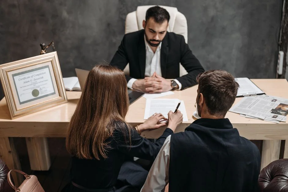 Zwei Personen sitzen einem Berater am Schreibtisch gegenüber und unterschreiben ein Dokument