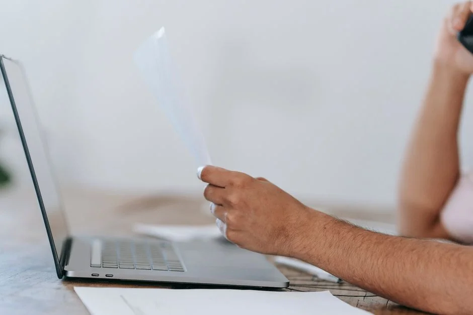 Person liest ein Dokument neben einem Laptop und telefoniert